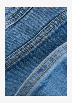 Denimová látka v odtieňoch modrej s viditeľným prešívaním v kontrastnej zlatej farbe, predstavujúca textúrovaný povrch a diagonálne záhyby.