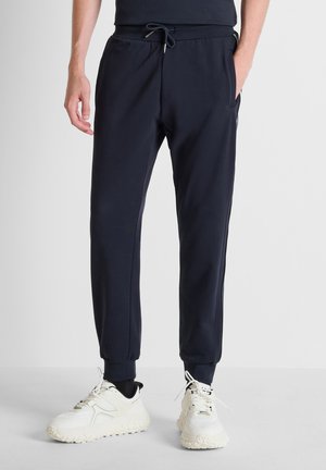 Uomo con pantaloni jogger neri con polsini elasticizzati e sneakers bianche chunky, in piedi contro uno sfondo chiaro uniforme.