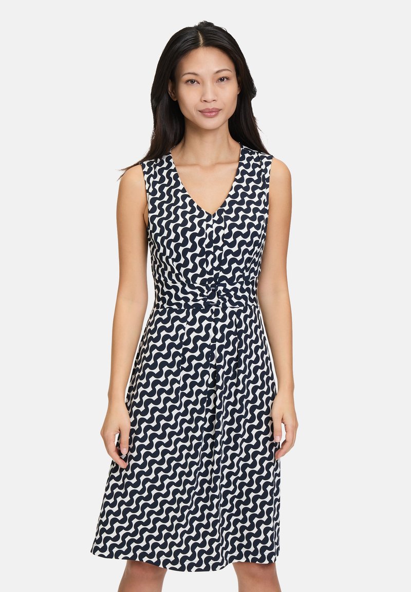 Ärmelloses Kleid in Marineblau mit weißem Wellenmuster, V-Ausschnitt-Design, Knoten-Detail an der Taille, knielang mit Knöpfen auf der Vorderseite.