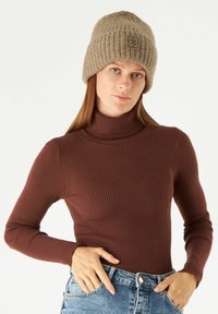 Brown ribbed turtleneck top paired with light blue denim jeans. Female model wears a knitted beige beanie with a small logo patch.