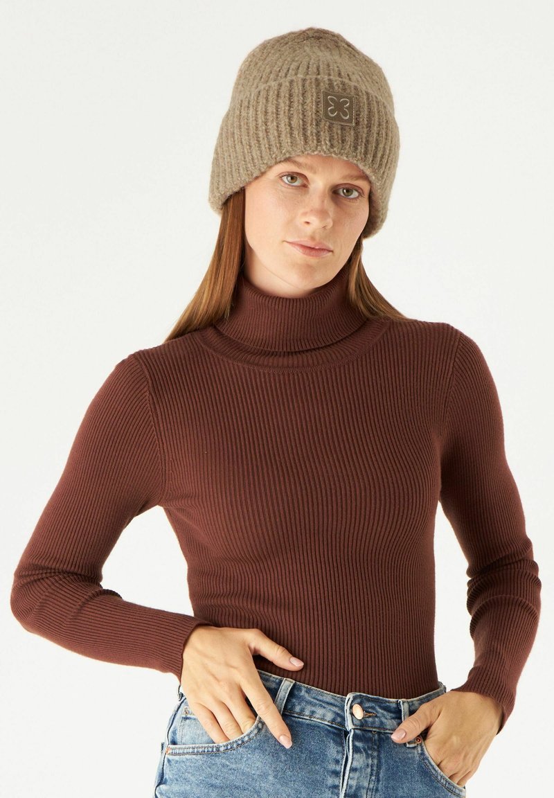 Brown ribbed turtleneck top paired with light blue denim jeans. Female model wears a knitted beige beanie with a small logo patch.