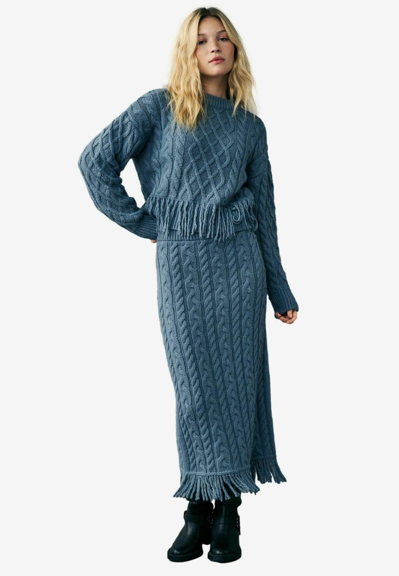 Set din două piese tricotat în albastru deschis, cu model de tricotare în cablu, cu tiv franjurat la pulover și la fustă, asortat cu botine negre până la glezne.