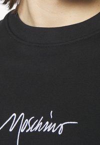 Gros plan d'un t-shirt noir à col rond avec un texte en cursive blanche brodé près de la zone de la clavicule.