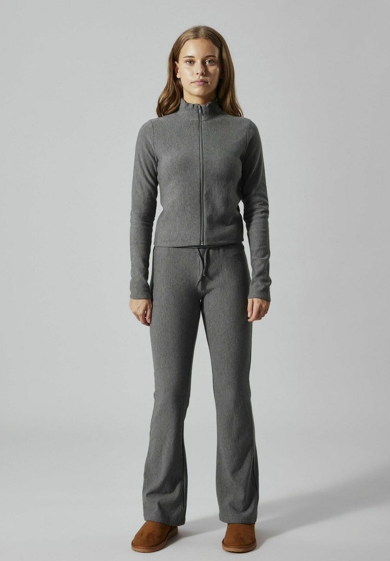 Veste zippée grise et pantalon évasé assorti en tissu doux et texturé. Le modèle porte des chaussures à enfiler marron.