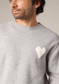 Pull gris clair avec un col côtelé, mettant en avant un détail en forme de cœur blanc sur la poitrine, fabriqué à partir d'un matériau doux et texturé.