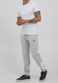 Weißes T-Shirt mit kurzen Ärmeln, graue Jogginghose mit schwarzen Akzenten und einem weißen Patch am Bein, kombiniert mit schwarzen Sneakers. Glatter Stoff, lässiges Design.