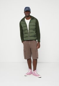 Roheline patjadega vest koos kootud käistega valge T-särgi peal, kombineeritud pruunide cargo-lühikesed püksid ja roosad kõrged tossud. Sinine ruuduline müts.