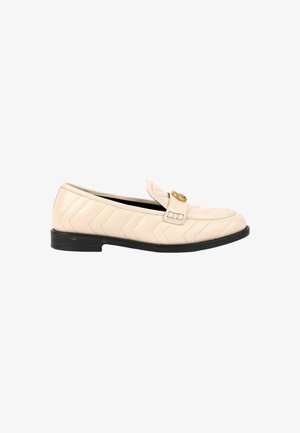 Mocassin en cuir beige avec matelassage chevron, semelle basse noire et boucle circulaire dorée sur le dessus.