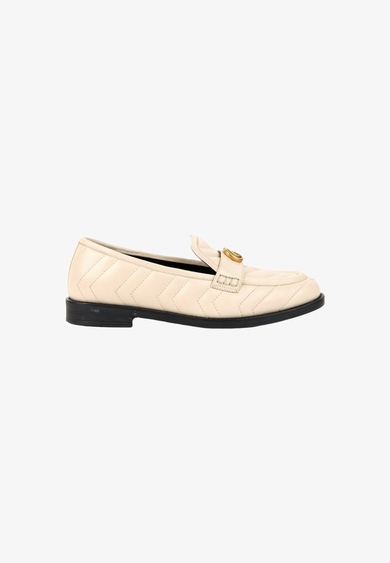 Mocassin en cuir beige avec matelassage chevron, semelle basse noire et boucle circulaire dorée sur le dessus.