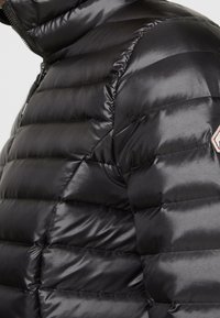 Sort dunjakke lavet af skinnende, quiltet materiale. Har en høj krave og lynlås i siden. Rødt og hvidt diamantemblem på overarmen.