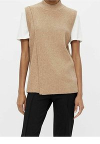 Une personne porte un gilet tricoté sans manches beige superposé à une chemise blanche à manches courtes, associée à un pantalon noir, sur un fond uni.