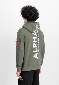 Sudadera verde oliva con un gran logo blanco "ALPHA INDUSTRIES" en la parte posterior, con un bolsillo tipo canguro en el frente y un bolsillo en la parte lateral del brazo.