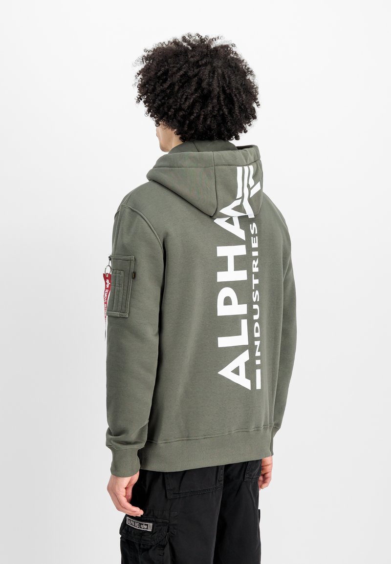 Sudadera verde oliva con un gran logo blanco "ALPHA INDUSTRIES" en la parte posterior, con un bolsillo tipo canguro en el frente y un bolsillo en la parte lateral del brazo.
