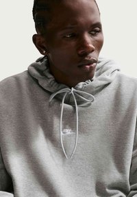 Homme portant un sweat à capuche Nike gris avec des cordons, regardant légèrement vers la droite, sur un fond clair uni.