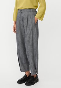 Szare spodnie w kratę z plisami, bocznymi kieszeniami i mankietami zapinanymi na guziki, zestawione z czarnymi mokasynami. Materiał wydaje się mieć strukturę i być dopasowany.