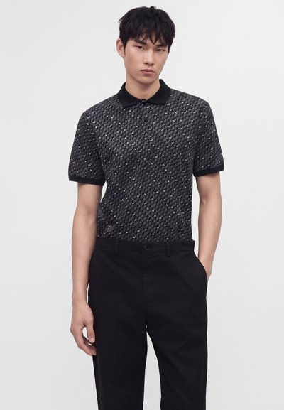 Jeune homme portant un polo noir à motif avec col et un pantalon noir, debout avec une main dans la poche sur un fond uni.