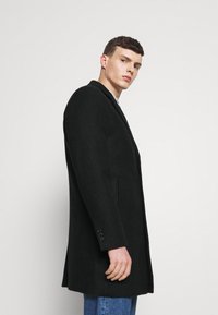 Abrigo de mezcla de lana negro con un patrón texturizado, diseño de un solo botón, cuello con muesca y bolsillos laterales. Cuenta con puños de cinco botones.