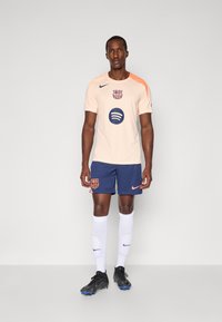 Peach Nike football jersey with navy shorts, featuring Barcelona logo, Spotify graphic, and orange accents. Worn with white socks and black cleats.