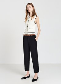 Top bianco senza maniche con bordi neri e bottoni, abbinato a pantaloni neri a tre quarti e una cintura intrecciata marrone. Ballerine nere a punta completano il look.