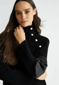 Maglione nero a coste con colletto alto, caratterizzato da bottoni in argento e dettagli in tessuto a contrasto sulle maniche. Texture morbida e design aderente.