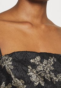 Haut noir brodé avec motif floral doré, avec tissu texturé et fines bretelles. Vue rapprochée du col et de la zone des épaules.