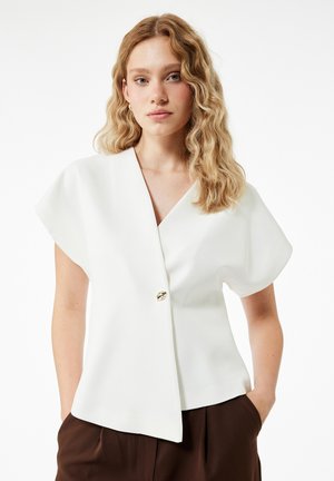 White short-sleeve blouse with a v-neck, featuring a gold button accent and a structured design. Smooth texture, paired with brown trousers.