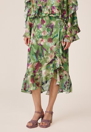 Donna che indossa una gonna midi verde a fiori con volant e orlo asimmetrico e sandali mauve in pelle scamosciata con tacco a blocco e cinturini alla caviglia.