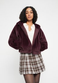 Naine kannab burgundia plush-jope, mille all on valge kootud sviiter ja pruun ruuduline plisseeritud seelik, seistes ühtlase valge tausta ees.