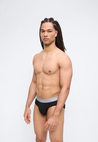 Fit moški z dolgimi dreadlocki, oblečen v črne Calvin Klein spodnje hlače, stoji ob preprostem svetlem ozadju in je rahlo obrnjen proti levi.