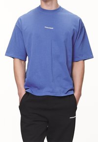 Camiseta azul de manga corta hecha de algodón con cuello redondo. Presenta un logo blanco "PEGADOR" en el pecho. Combinada con pantalones negros.