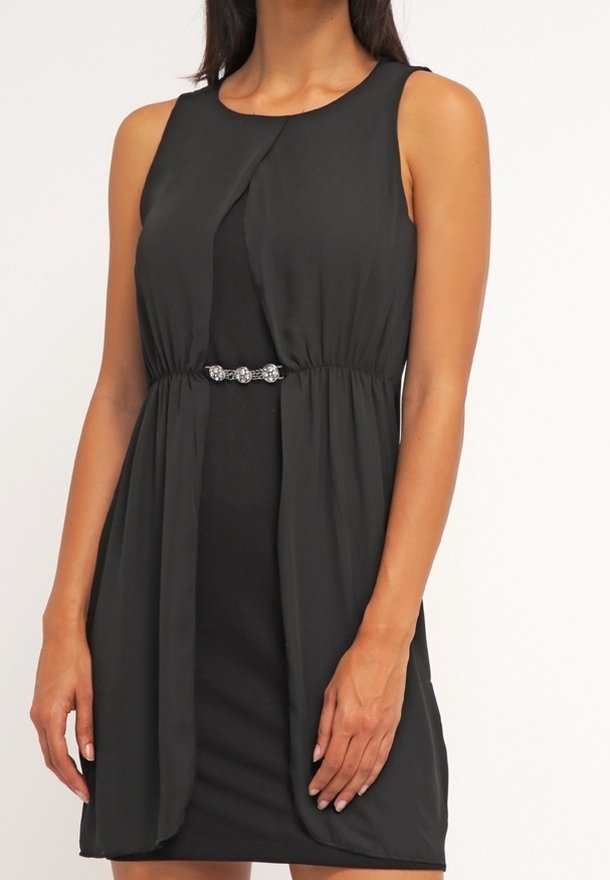 Robe noire sans manches avec une taille froncée et un fermoir décoratif orné de bijoux au centre, portée par une personne debout sur un fond uni.
