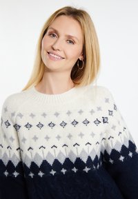 Pīnēts džemperis ar krēmkrāsas ķermeni, zilām un pelēkām ģeometriskām rakstām, ribu apkakli un teksturētām tumši zila krāsas piedurknēm.
