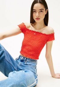 Top rosso scollato con arricciature testurizzate e orli a volant, con un piccolo logo. Abbinato a jeans in denim blu a vita alta.