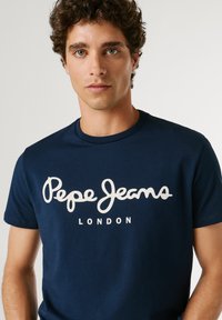 Junger Mann mit lockigem Haar, der ein dunkelblaues T-Shirt mit weißem "Pepe Jeans London"-Logo vor schlichtem hellem Hintergrund trägt.