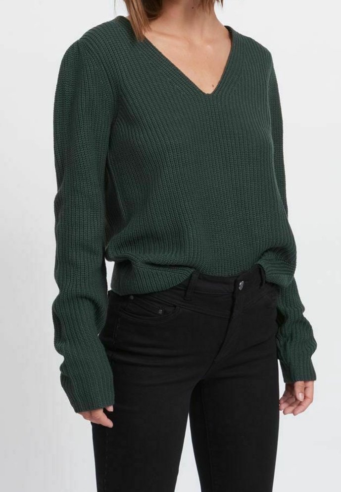 Ciemnozielony sweter w serek, wykonany na drutach, z długimi rękawami. Teksturowany materiał, luźny krój, noszony na czarnych spodniach. Brak wyraźnych wzorów lub akcentów.