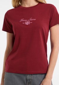 Burgundská bavlnená tričko s krátkymi rukávmi, s jemne ružovým vyšitým logom a emblémom "Guess Jeans" na prednej strane.