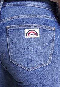 Jean en denim bleu avec une poche arrière ornée d'un patch logo Wrangler blanc et des coutures superposées formant un motif en chevron.