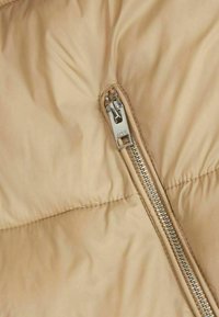 Beige pufferjacka i tyg med en texturerad yta; har en silvrig dragkedja längs sidan för stängning och detaljer av söm.