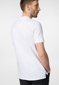 Biała koszulka polo z krótkim rękawem, wykonana z bawełny, klasyczny kołnierzyk, minimalistyczny design z małym czerwonym logo na górnej części pleców oraz wentylacją po bokach.