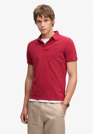 Ung mand iført en rød kortærmet polo, beige bukser og et armbåndsur med læderrem, står med den ene hånd i lommen.