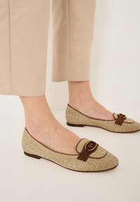 Ballerines tissées beiges avec un bout rond, comprenant un accent en cuir marron et un détail de boucle. Les semelles sont plates et lisses.