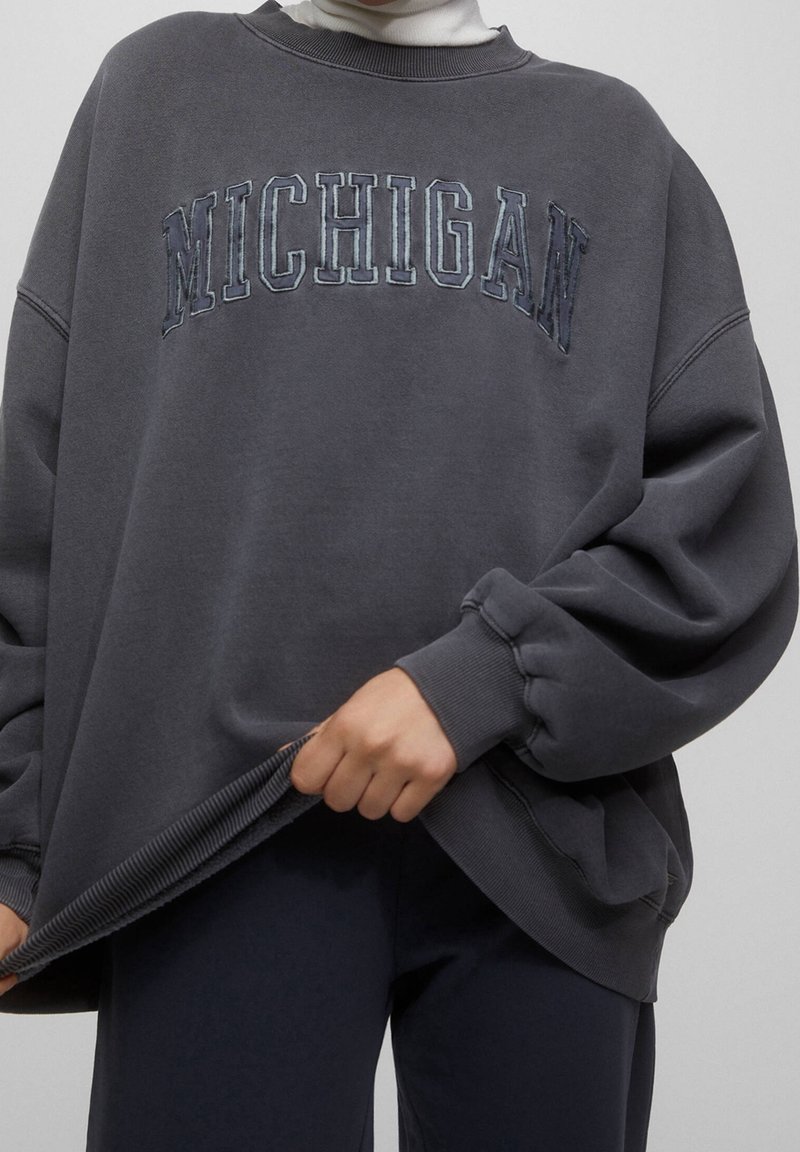 Personne portant un sweat-shirt oversize gris foncé avec "MICHIGAN" brodé sur la poitrine, tenant l'ourlet, visible du cou aux hanches.