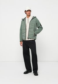 Doudoune vert clair avec un motif hexagonal texturé, accents noirs, fermeture éclair intégrale, associée à un polo crème, un pantalon sombre et des chaussures noires.