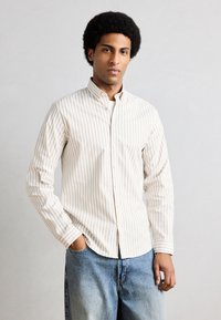 Homme avec une coiffure afro portant une chemise à rayures beige et blanche boutonnée et un jean bleu, debout avec une main dans la poche devant un fond uni.