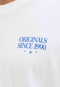 Camiseta de algodón blanca con texto azul que dice "ORIGINALS SINCE 1990" con acentos de estrellas debajo. Diseño clásico de cuello redondo.