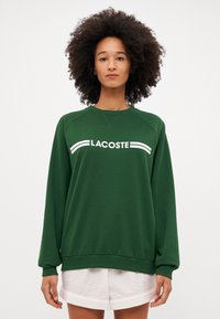 Groene sweatshirts met ronde hals, geribbelde boorden en zoom. Voorzien van een witte "LACOSTE"-logo aan de voorkant in een sportieve stijl.