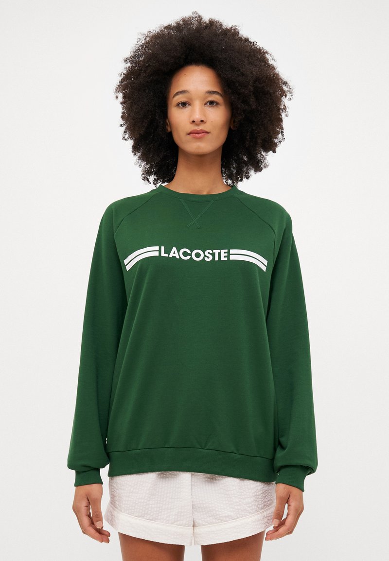 Groene sweatshirts met ronde hals, geribbelde boorden en zoom. Voorzien van een witte "LACOSTE"-logo aan de voorkant in een sportieve stijl.