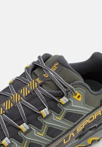 Grijze en olijfgroene veterschoenen voor trailrunning hebben een gestructureerde mesh bovenkant, gele accenten en een robuuste zool ontworpen voor grip.