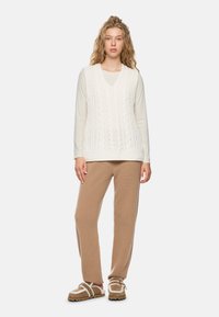 Woman with wavy blonde hair wearing a white long-sleeve top, sleeveless cable-knit sweater, beige knit pants, and shearling-lined slippers.