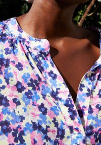 Kukallinen pusero kevyestä kankaasta, jossa on vaaleanpunaisia, sinisiä ja purppuran värisiä kukkia keltaisella taustalla, sekä V-mallinen pääntie nappilistan kanssa.